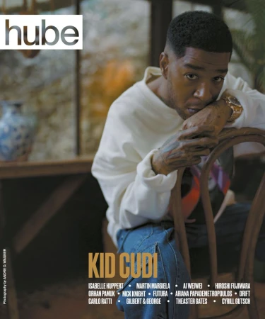 Hube magazine cover featuring Kid Cudi. Medium shot of an adult male in a white sweatshirt and jeans, sitting on a wooden chair.