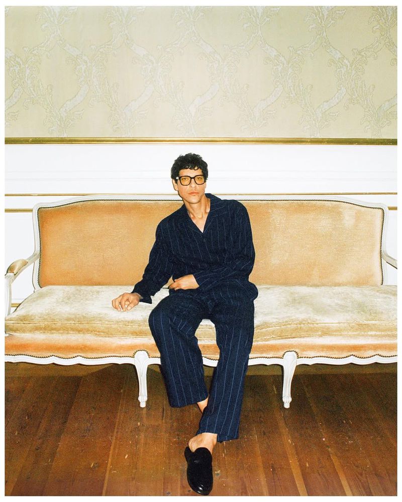 A man in a blue striped pajama suit sits on a vintage peach-colored couch in a room with patterned wallpaper and a wood floor.