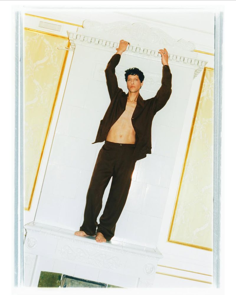 Young man in brown suit posing indoors with hands on the white ornate molding above, looking straight at the camera with a neutral expression.
