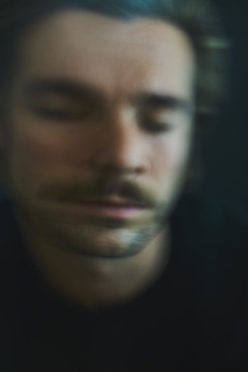 A blurry, dark portrait of a man with closed eyes, a mustache, and dark hair, set against a dark backdrop.
