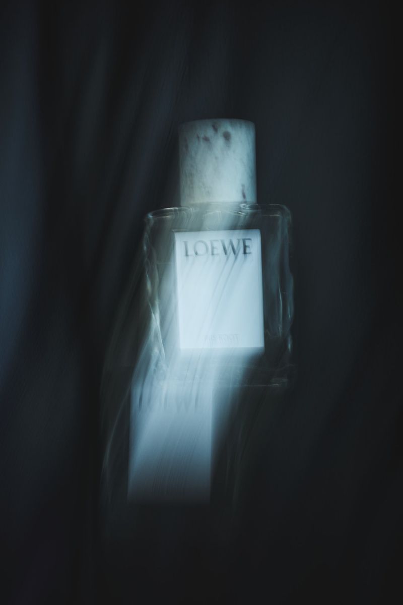 Close-up studio shot of a Loewe perfume bottle featuring clear glass, a white label, and a light blue background with blurred fabric.