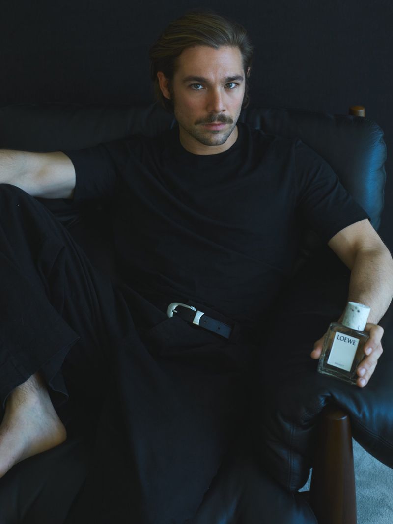 Adult male sitting in a leather chair, holding a Loewe perfume bottle in an indoor studio with a dark background.