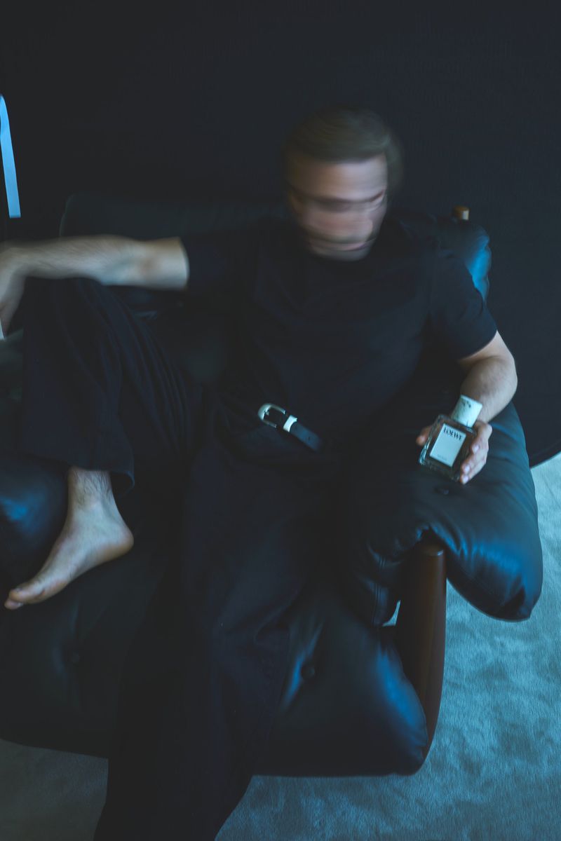 A man in dark clothing lounges on a leather chair holding a bottle, captured in a blurred, dynamic, and visually interesting composition.