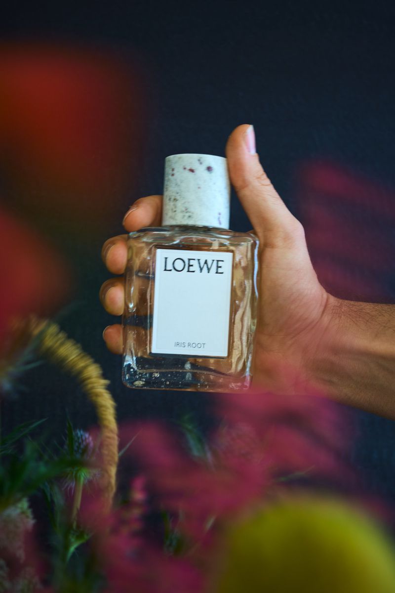 Clear Loewe Iris Root perfume bottle being held by a hand against a blurred, colourful floral background in a close-up shot.