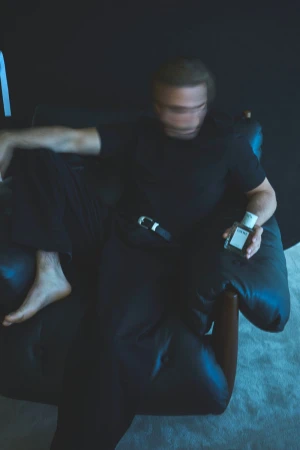 Carlos Cuevas seated barefoot in a black armchair holding a Loewe Esencia fragrance bottle with motion-blurred face.