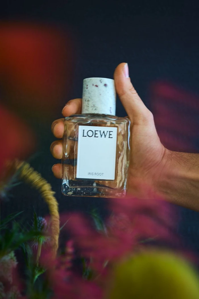 Hand holding a clear glass Loewe perfume bottle with a patterned cap against a blurred floral background