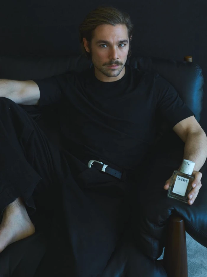 Actor Carlos Cuevas seated in a black leather armchair holding a bottle of Loewe perfume against a dark studio backdrop.