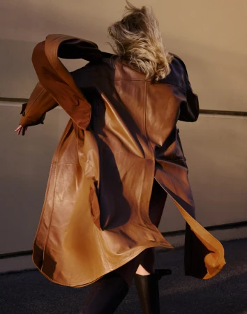 A woman with blonde hair is facing away from the camera wearing a long brown leather coat and black boots against a muted background.