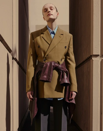 Fashionable young woman in a tan jacket and leather wrap stands outdoors in sunlight, presenting modern style and urban aesthetic.