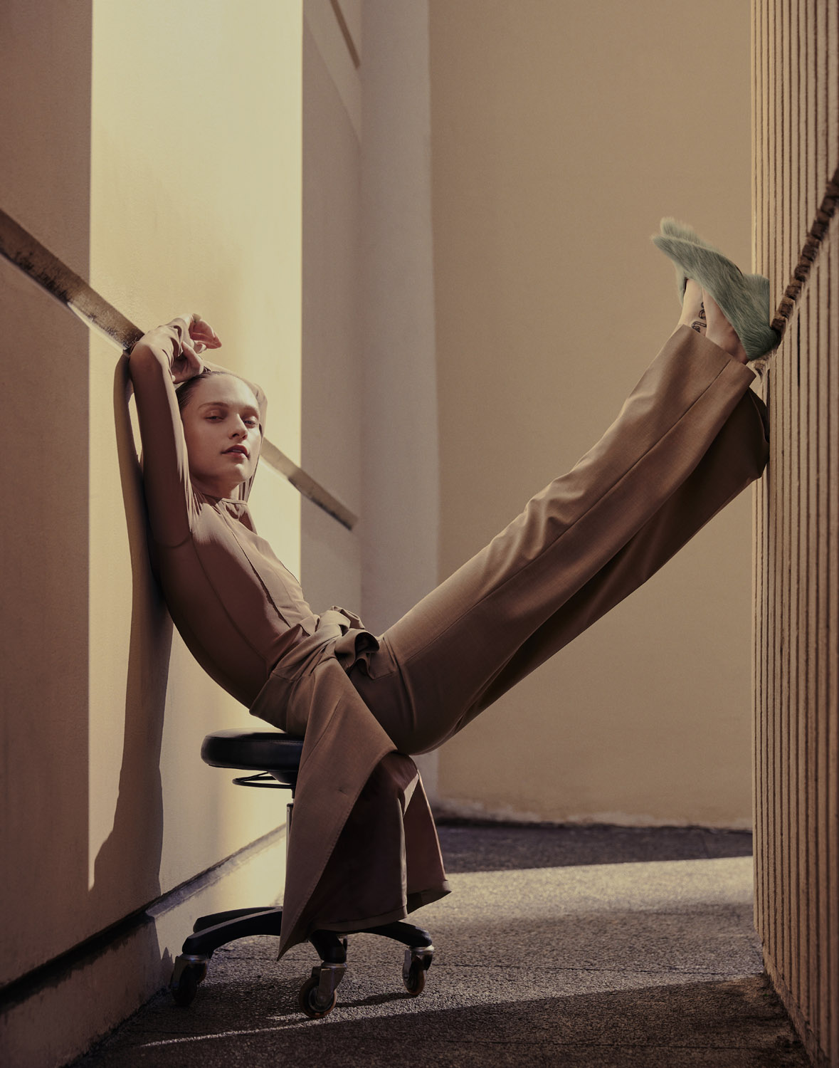 A fashionable woman in a brown suit and green shoes leans against a wall on a rolling office chair, looking directly at the camera.