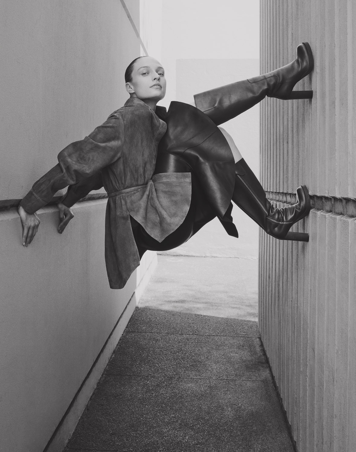 Monochrome fashion shot of an adult woman in a coat and high-heeled boots posing outdoors against a wall with an angular composition.