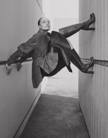 Monochrome fashion shot of an adult woman in a coat and high-heeled boots posing outdoors against a wall with an angular composition.