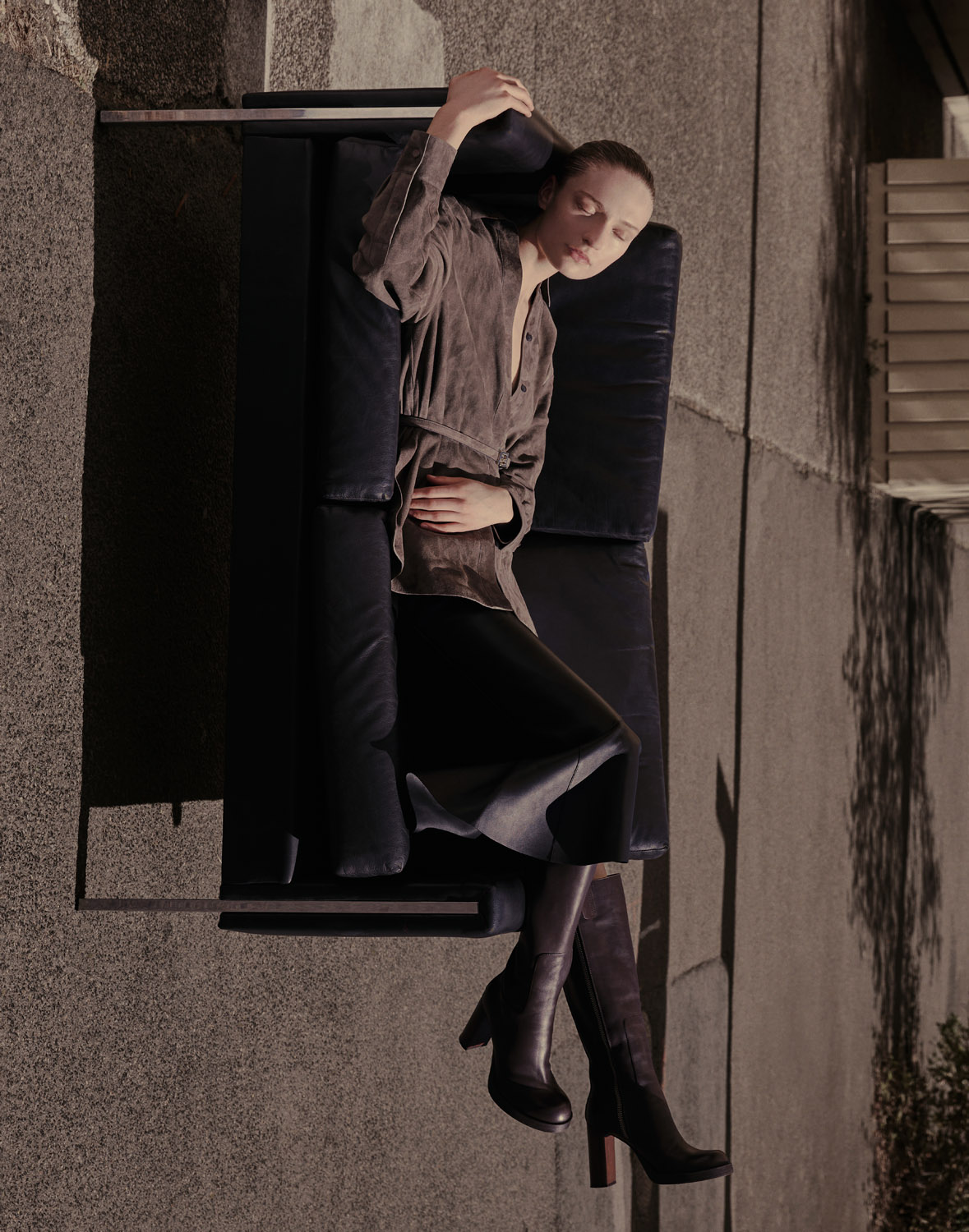 An adult female model reclines on a black chaise lounge wearing a tan blouse, black skirt, and knee-high brown leather boots indoors.