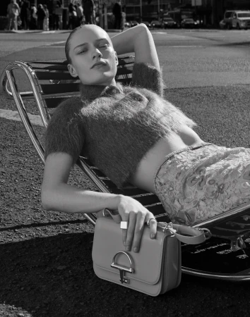Black and white fashion shot of a woman reclining on a chair outdoors with a handbag, wearing a crop top and printed shorts.