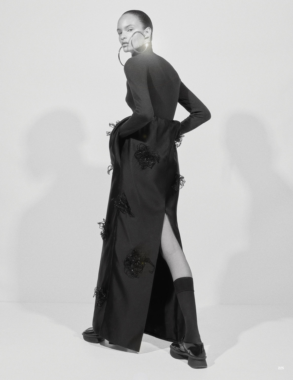 Elegant woman in a black gown and large hoop earrings poses in a studio, showcasing fashion and editorial style against a plain background.