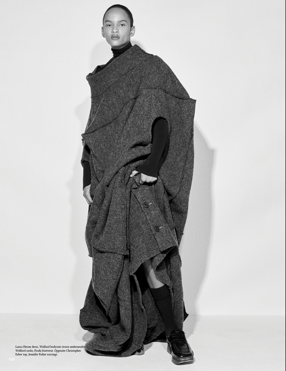 Fashion shot of a young adult woman in a grey tweed coat with a black turtleneck, socks, and shiny black shoes in a photo studio.