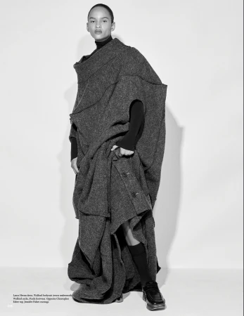 Fashion shot of a young adult woman in a grey tweed coat with a black turtleneck, socks, and shiny black shoes in a photo studio.