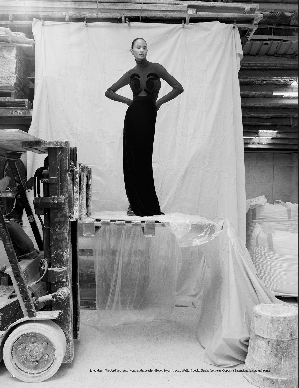 An adult female fashion model poses in a long, dark gown while standing atop a pallet in what appears to be a warehouse.