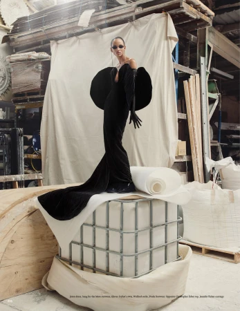 Elegant woman in a black gown with statement shoulders stands atop a metal structure indoors with white fabric backdrop in a fashion editorial.