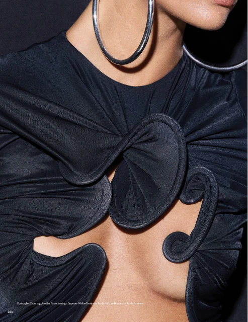 Close-up of a woman wearing a black top with a structural design and large silver hoop earrings against a dark background.