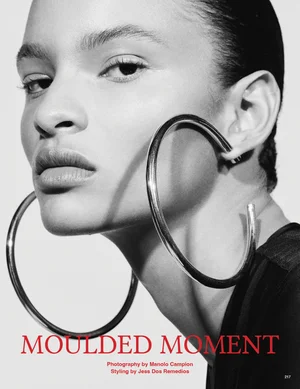 Close-up portrait of a woman wearing oversized metallic hoop earrings and a dark top against a plain light background.