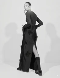 Model posing in a long black dress with embellishments, dark socks, platform shoes, and large hoop earrings in a monochrome studio.