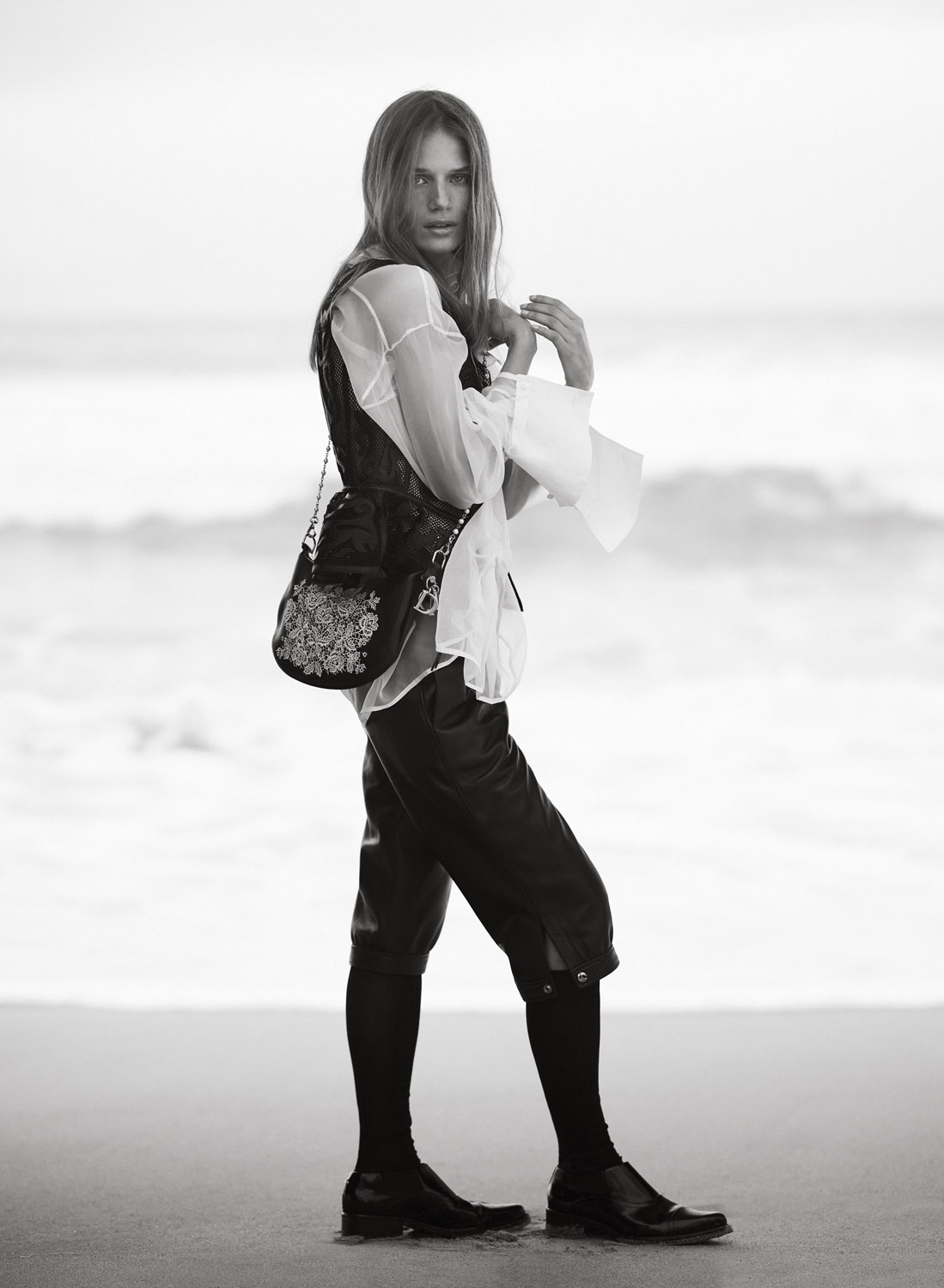 Black-and-white fashion shot of a woman in vintage-style clothing standing on a beach, wearing a vest, blouse, and culottes.