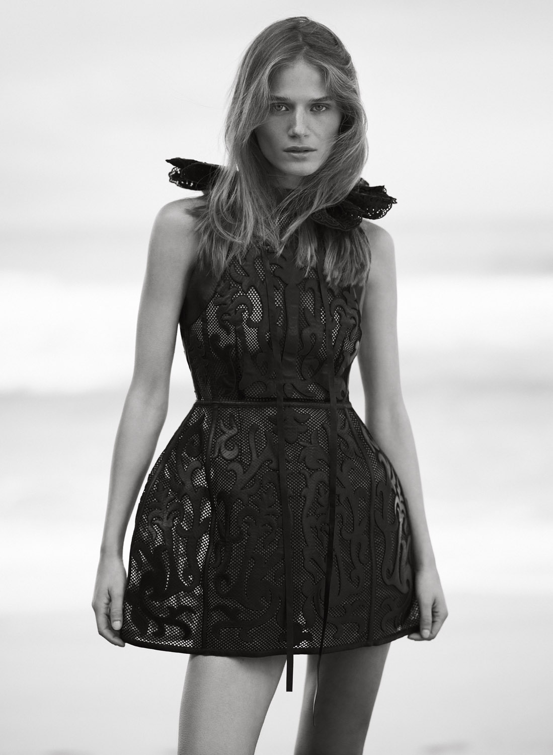 Monochrome portrait of a young woman in a black patterned dress with a ruffled collar, standing outdoors with a bright, neutral background.