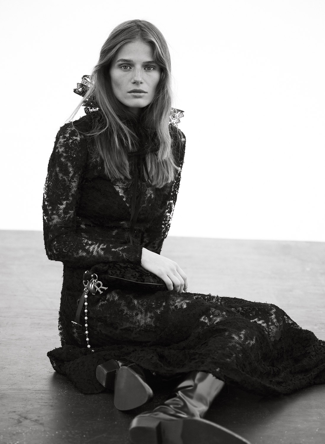 Elegant portrait of a woman in a long black lace dress, seated indoors against a white backdrop with dark flooring.