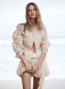 Model wearing a sheer cream lace jacket and matching shorts posing outdoors against an ocean beach backdrop.