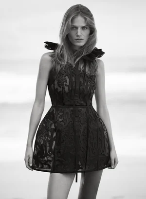 Model in a black lace patterned dress with ruffled collar standing against an outdoor coastal background