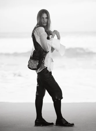 Model in a sheer white blouse and black cropped trousers holding a shoulder bag while standing on a beach in black and white.