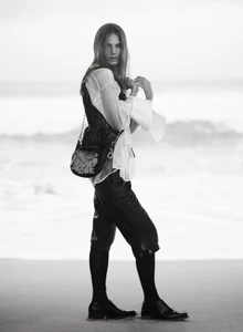 Model in a sheer white blouse and black cropped trousers holding a shoulder bag while standing on a beach in black and white.