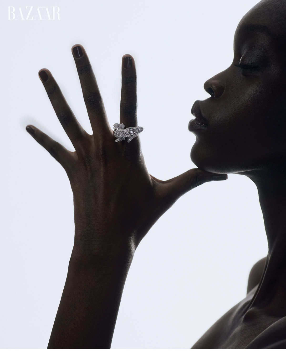 High-contrast fashion shot of an adult woman wearing a gemstone ring, her hand near her face, with white gradient background and 'BAZAAR' logo.