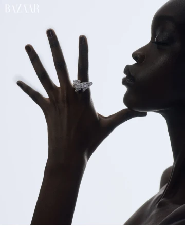 High-contrast fashion shot of an adult woman wearing a gemstone ring, her hand near her face, with white gradient background and 'BAZAAR' logo.