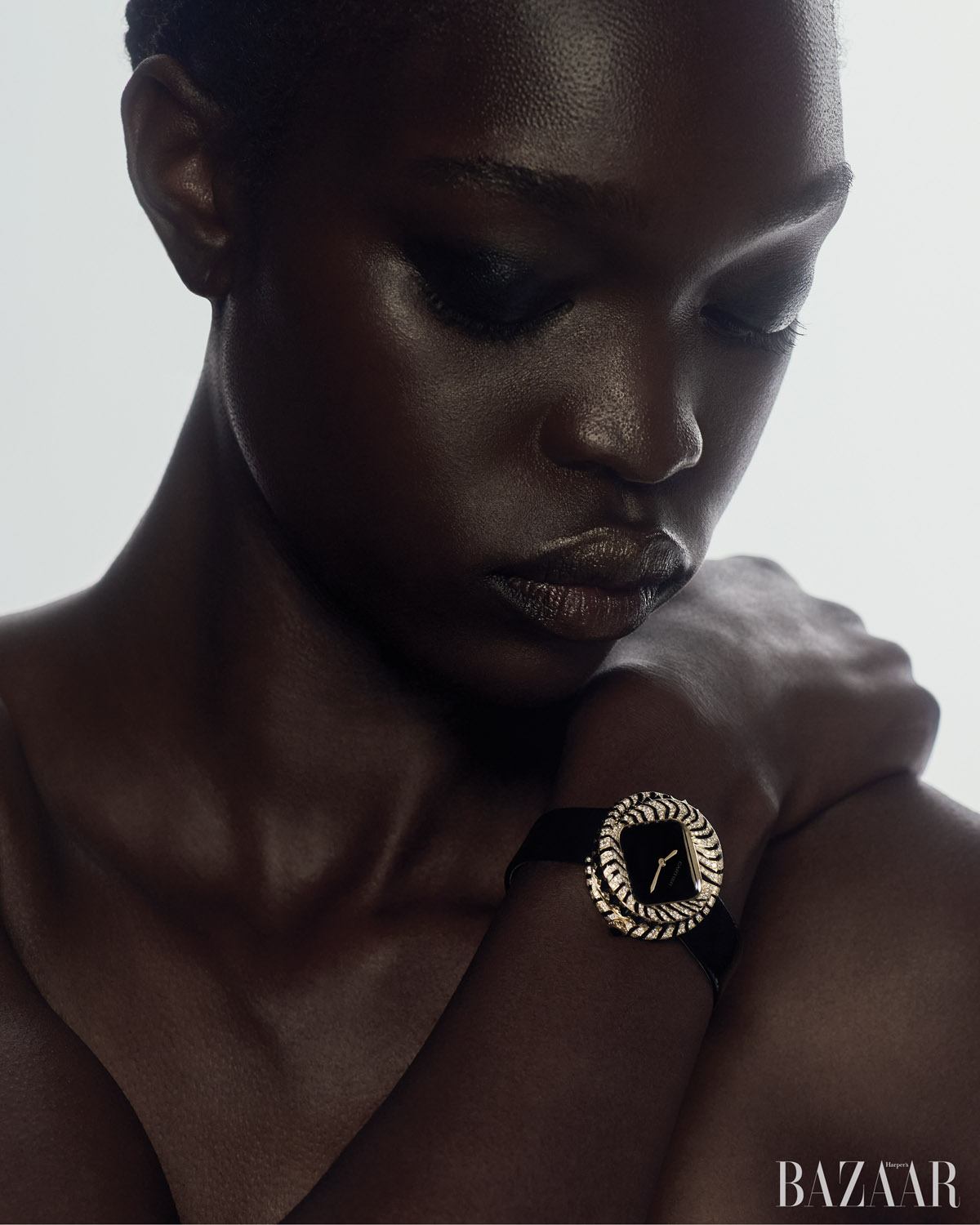 Close-up studio shot of a Black adult female fashion model wearing a black wristwatch with an ornate gold bezel and looking down.