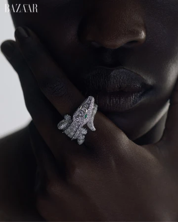 Close-up of a dark-skinned person's face and hands, showcasing a detailed crocodile-shaped ring adorned with diamonds, emphasizing luxury jewelry.