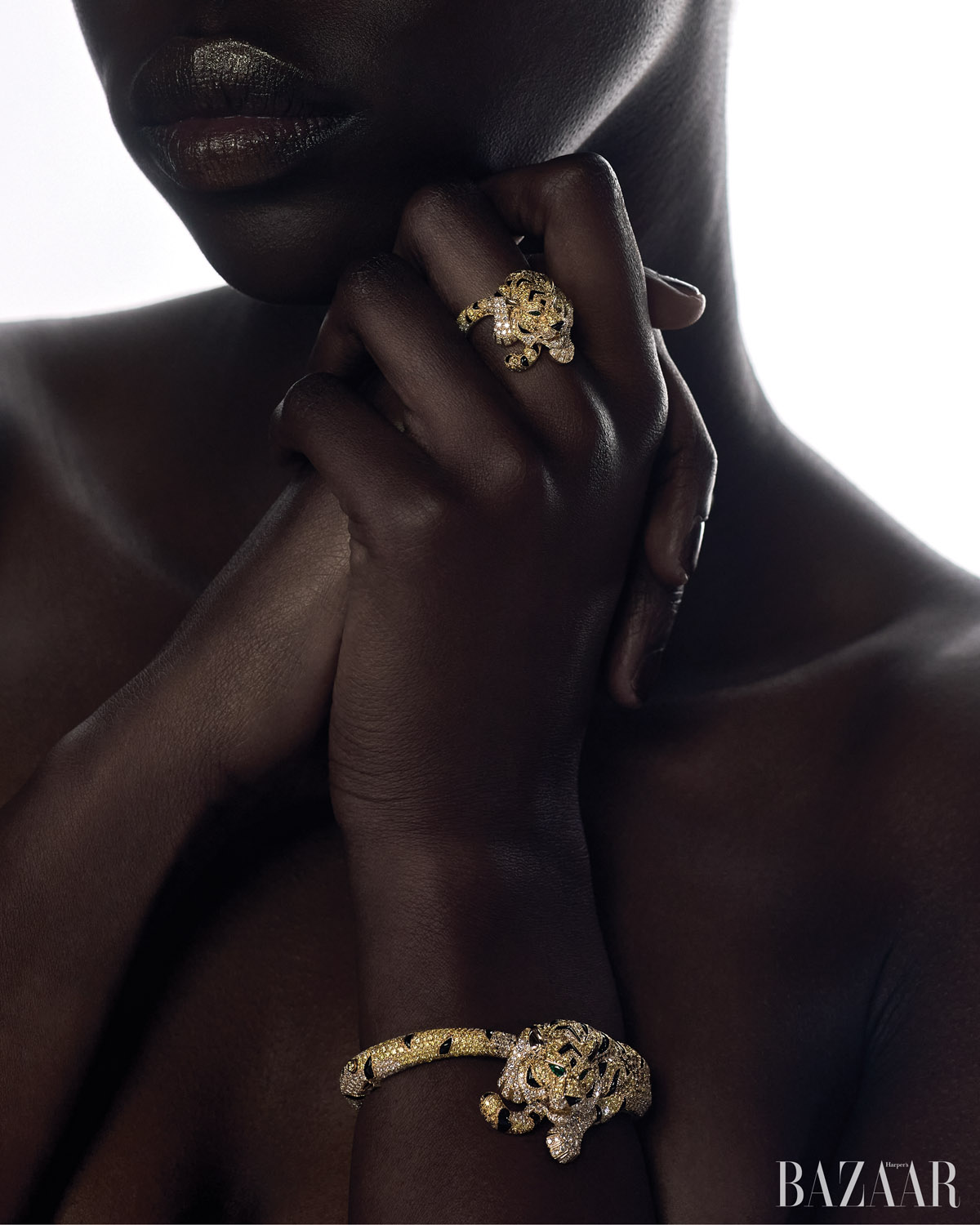 Dark-skinned model poses in a close-up studio shot, showcasing gold tiger jewelry: a ring and a bracelet, with a Bazaar logo visible.