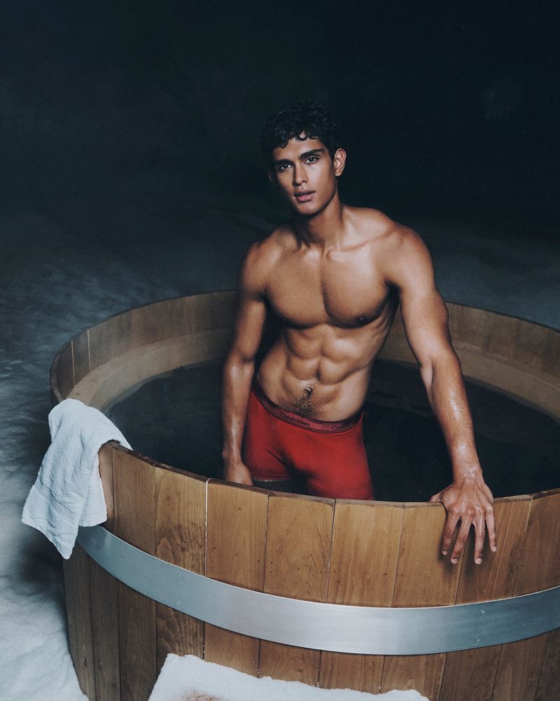 Man in red shorts in a wooden tub. Athletic build, dark curly hair. Towel on the tub edge. Moody lighting. Fitness, health, and personal care.