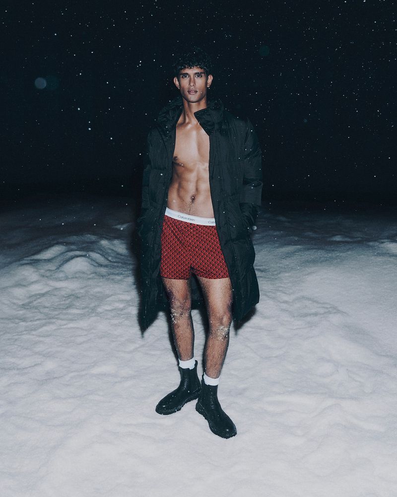 Male fashion model poses shirtless in boxer shorts and a long coat in a snowy, dark outdoor environment against a starry sky.