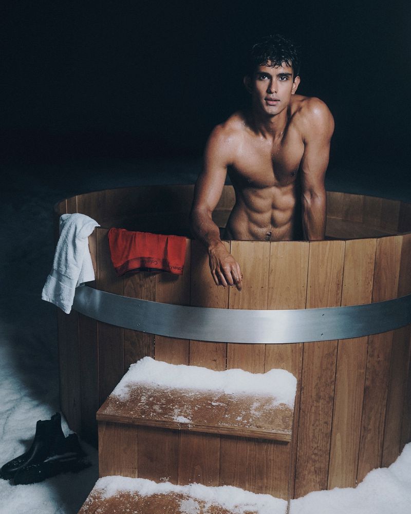 Shirtless male model poses in a wooden hot tub outdoors in snowy setting, creating a striking contrast. Fitness and wellness themes.
