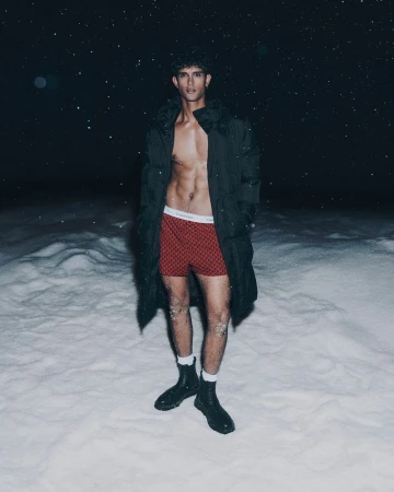Male model wearing red patterned boxers, white socks, black boots, and a black puffer coat standing outside in snow