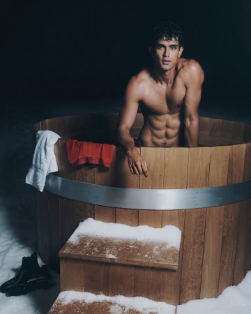 Shirtless male model leaning over a wooden hot tub outdoors in a snowy setting at night.