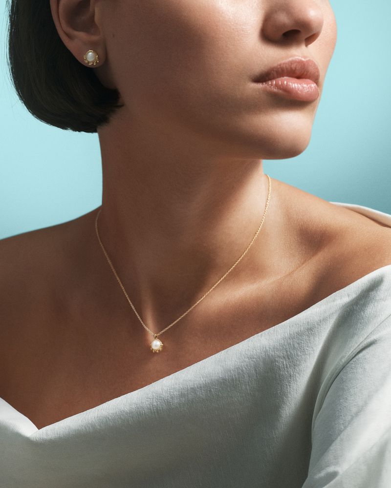 Close-up of a woman wearing a gold necklace and earring jewelry set with pearl accents against a gradient blue background.