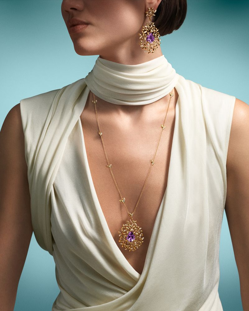Close-up of a woman wearing a white dress and elaborate gold jewelry with amethyst stones against a light blue background.