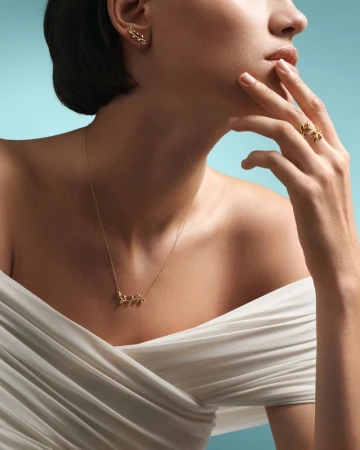 Adult woman poses wearing gold leaf earrings, a necklace and ring with an off-the-shoulder white dress on a light blue background.