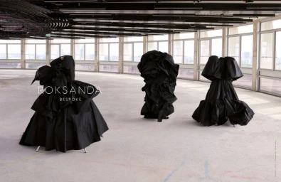 Three sculptural black voluminous gowns standing on display in a spacious empty loft interior with panoramic windows.