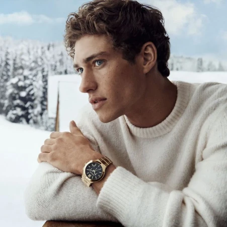 Handsome man in a white sweater and gold watch in a snowy outdoor landscape, portrait-style photograph showcasing fashion and style.