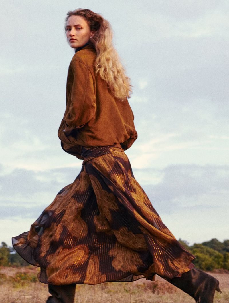 A woman with blonde hair, wearing a brown jacket and flowing floral skirt, walks through a field of tall grass under a cloudy sky.