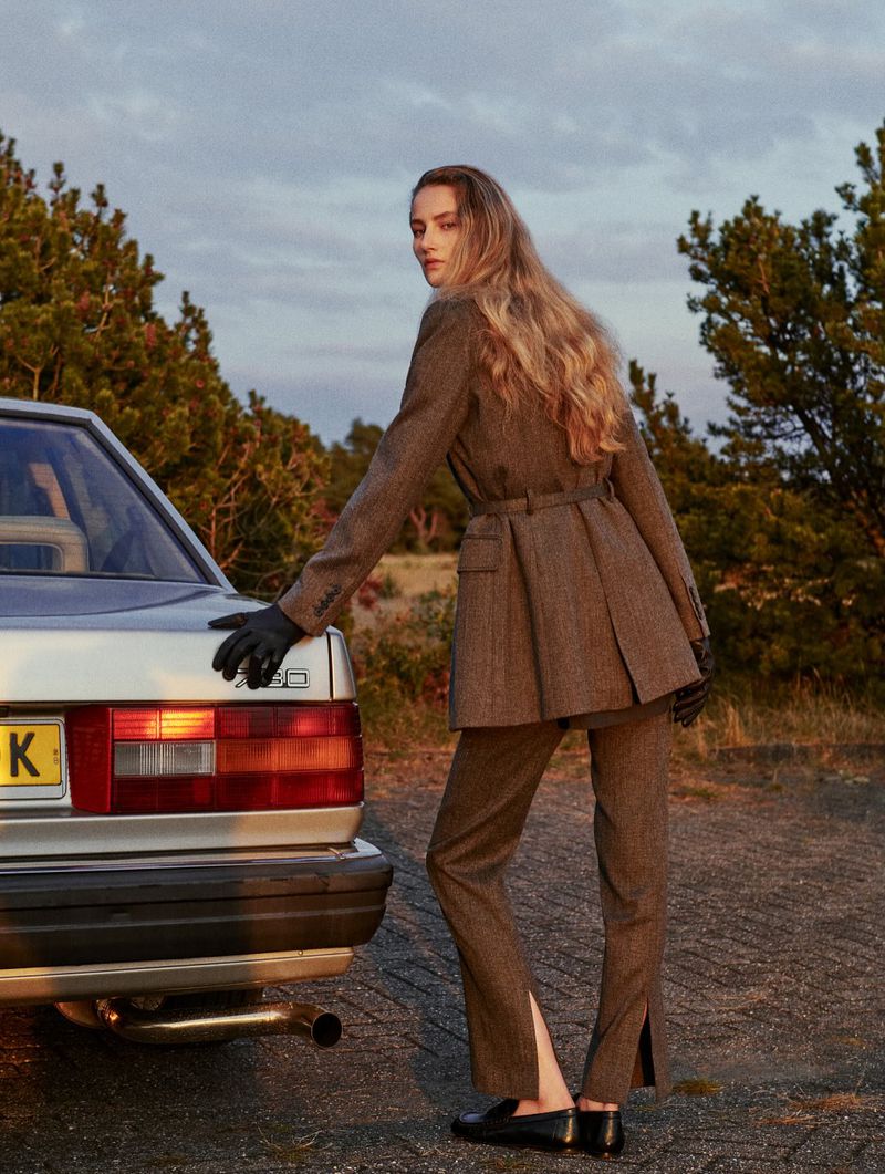 Woman in a brown suit and black gloves poses next to a silver car in an outdoor setting with green trees and a cloudy sky.